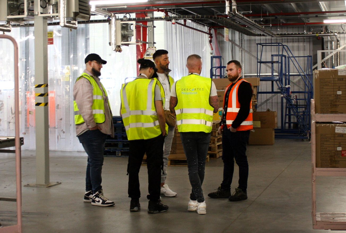 A group of employees wearing safety vests stand together in a warehouse. One of the safety vests is emblazoned with the Descartes logo on the back, representing how Descartes supports ecommerce operations and business growth.