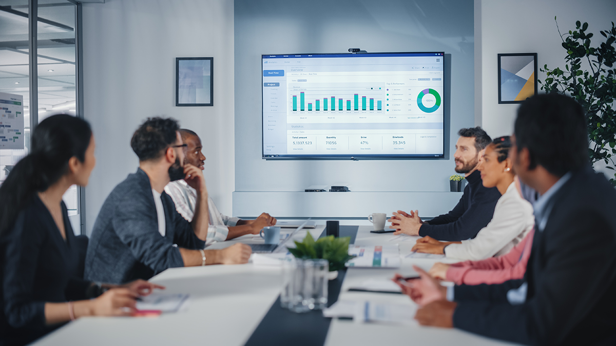 A group of ecommerce business leaders discusses adding multiple marketplaces while sitting at a conference room table, looking at a screen on the wall displaying business charts and graphs.