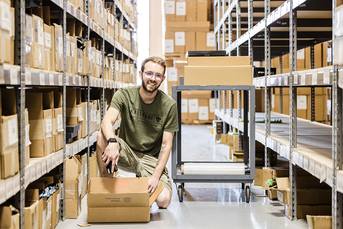 A worker kneels with a parcel and mobile device using multichannel ecommerce operations software in a warehouse aisle lined with shelves and boxes.