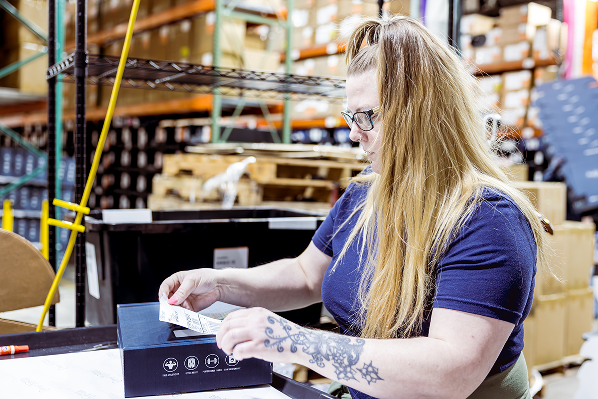 A warehouse worker stands at a packing station in a busy warehouse and affixes a shipping label to a parcel for multichannel ecommerce shipping.