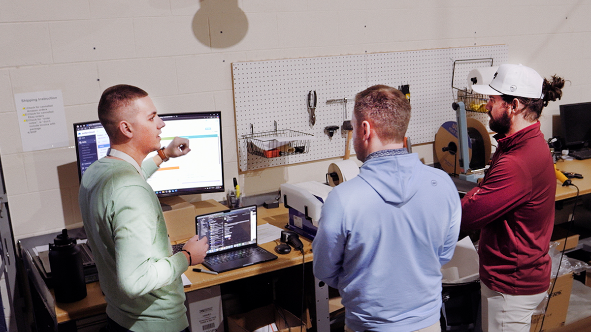 A warehouse manager stands at a shipping station with a laptop and large monitor, explaining shipping software for multichannel ecommerce to workers.