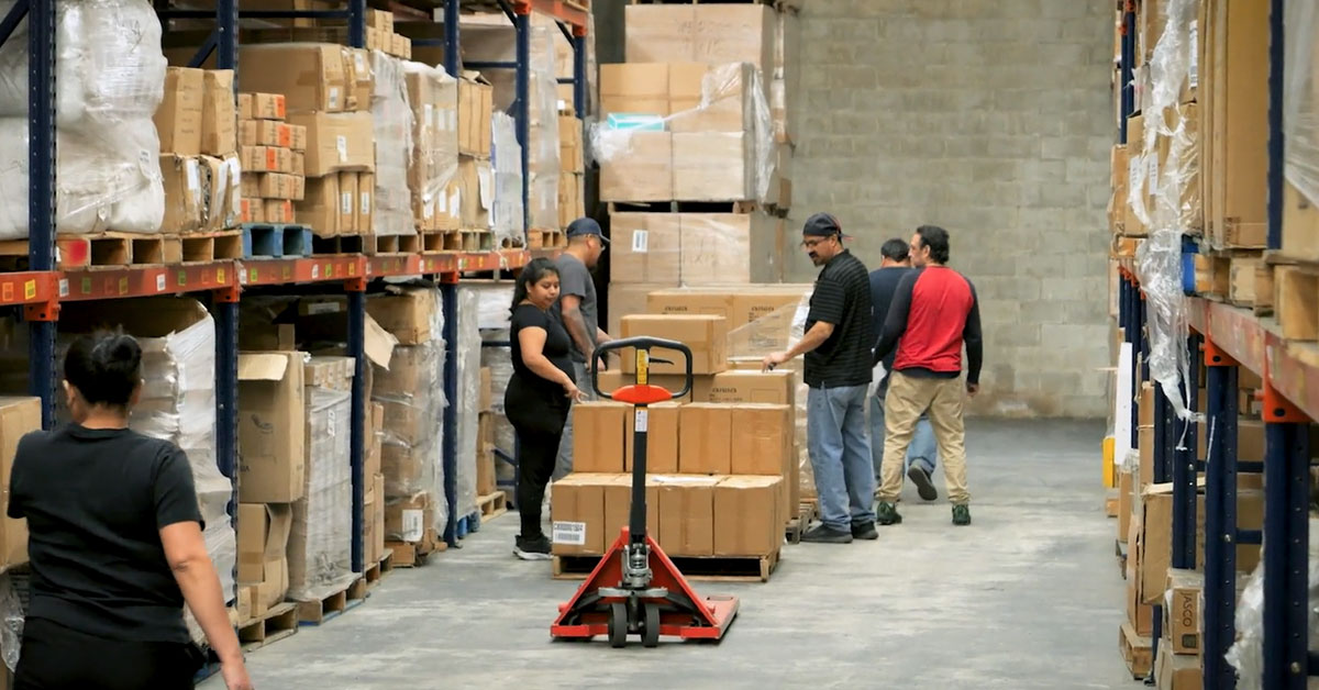 Warehouse team organizing and moving boxed inventory with a pallet jack between tall storage racks.