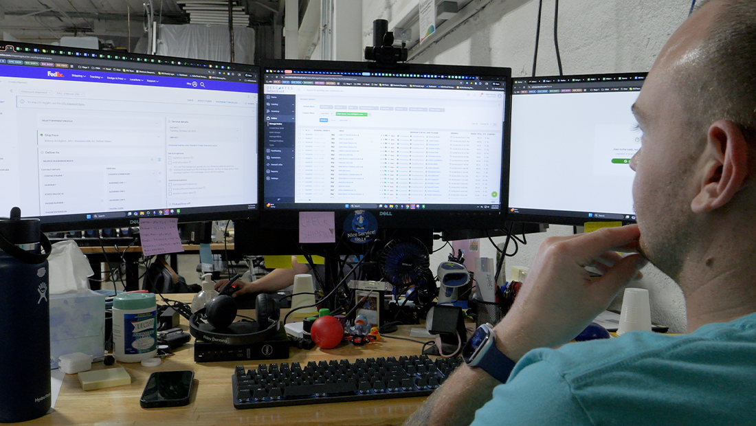 Employee reviewing shipping and order management software across three computer monitors at a warehouse workstation.