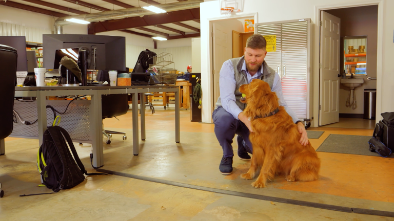 A man is petting a golden retriever dog in a warehouse office while a computer automatically creates, sends, and tracks purchase orders online.