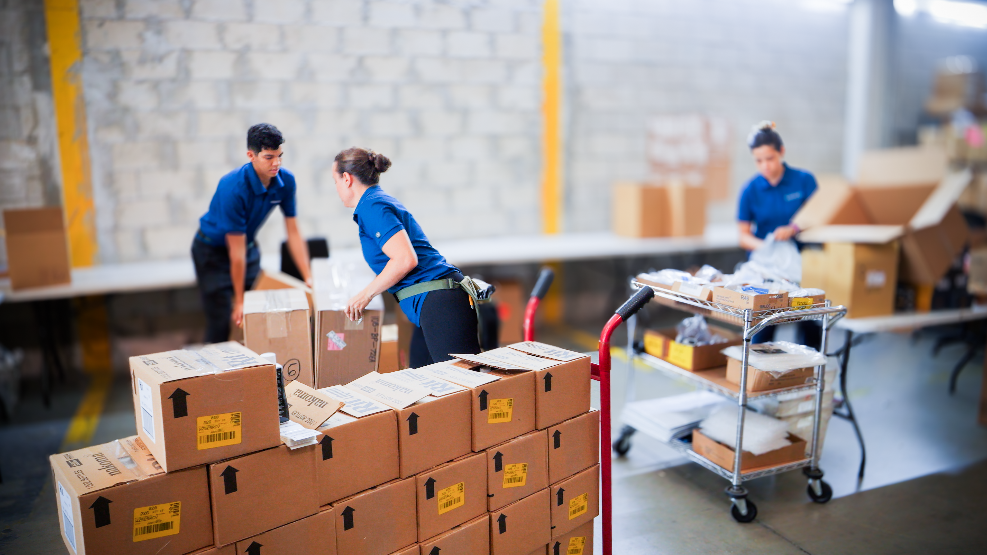 Three warehouse workers organize and move shipping boxes in a packing and fulfillment area and stacked cardboard boxes sit on pallets while employees lift packages and prepare items inside a large industrial warehouse.
