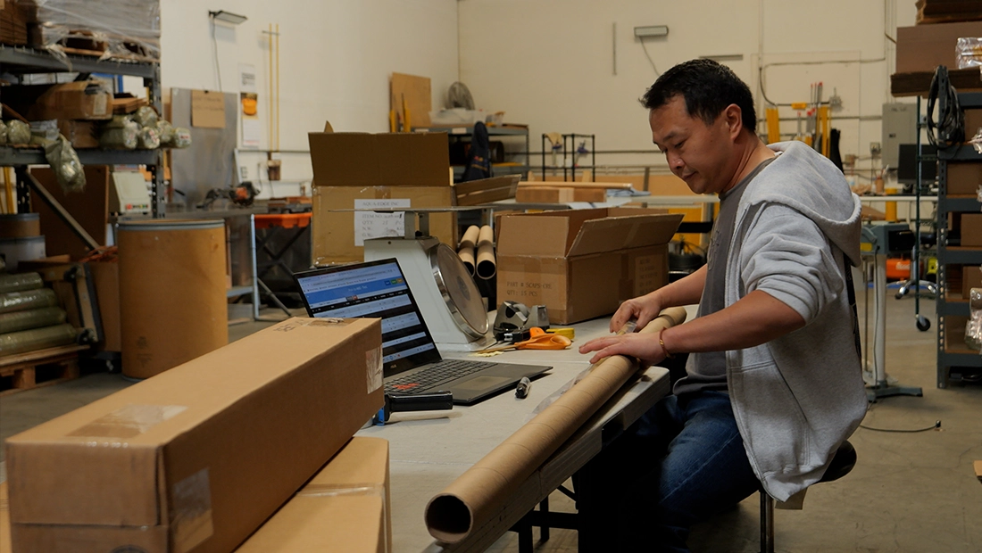 Warehouse worker packing items at a workstation with boxes, packing materials, and a laptop for order processing
