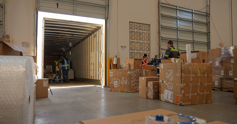 An image showing a warehouse docking bay. Goods are transferred from the back of a truck into a warehouse.