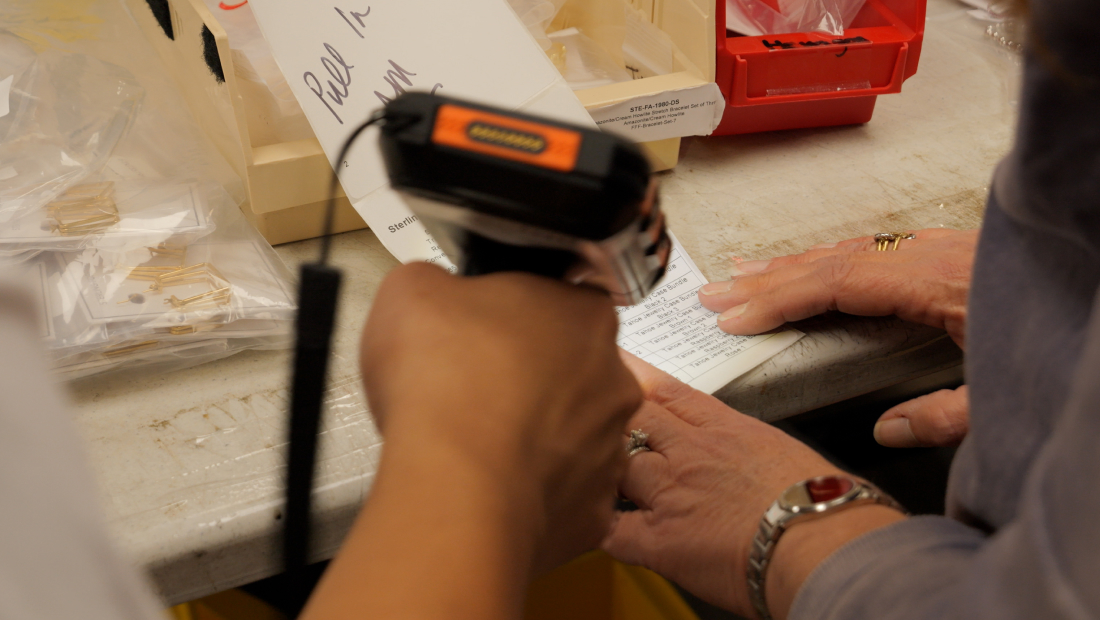 Hands using a handheld barcode scanner to scan a paper form on a workbench surrounded by small parts and storage bins.