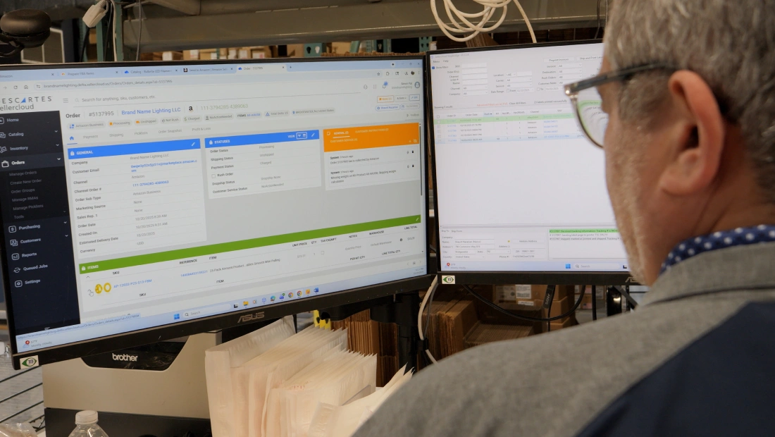 Warehouse manager reviewing order and inventory information on dual computer monitors displaying ecommerce and warehouse management software.