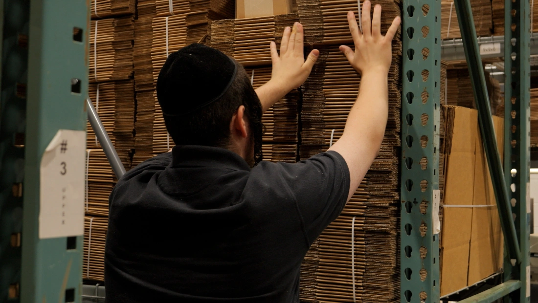 Warehouse employee organizing and handling stacks of flattened cardboard boxes on industrial storage shelving.