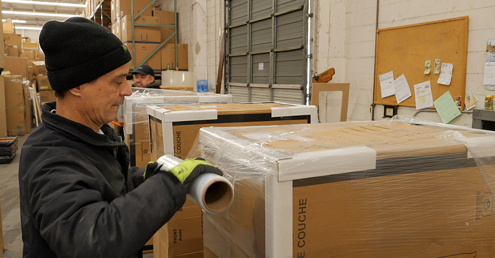 A warehouse worker wraps a large cardboard box with transparent plastic stretch wrap, with stacked boxes and another worker in the background.