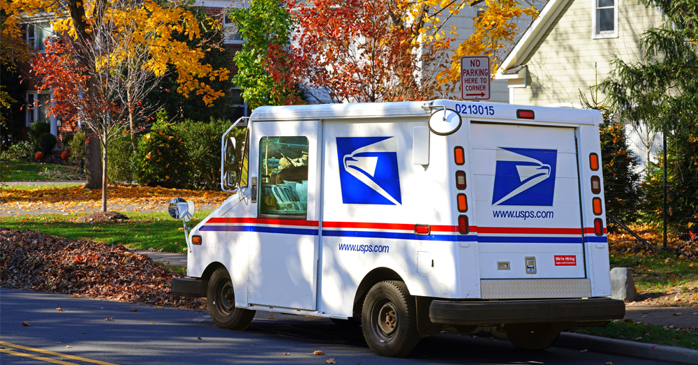 An image showing a USPS truck parked on a suburban street. Trees with red- and yellow-colored leaves suggest it is Fall.