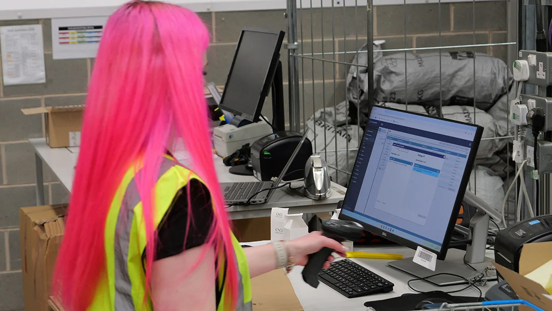Warehouse employee wearing a high-visibility vest using a handheld barcode scanner at a computer workstation.