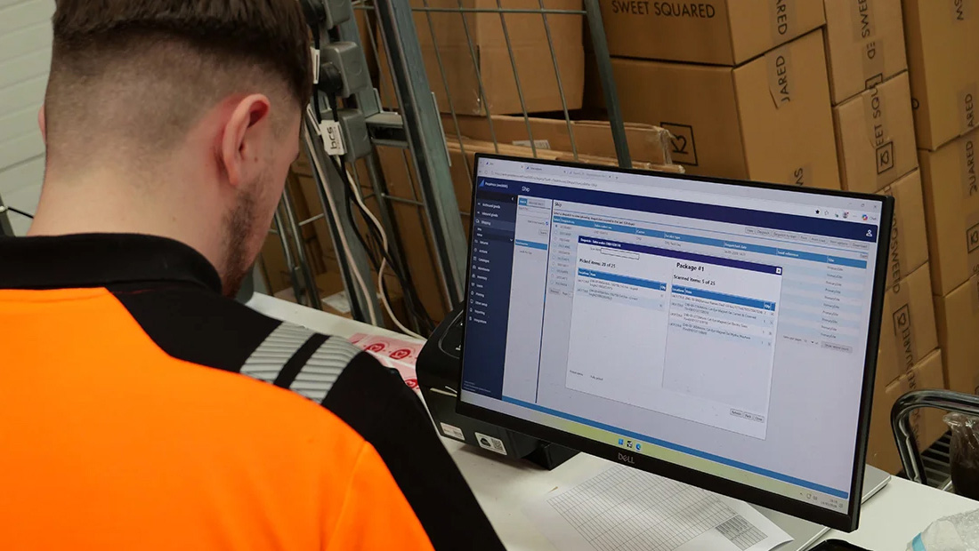 Warehouse employee using a computer-based warehouse management system at a packing station surrounded by Sweet Squared boxes.