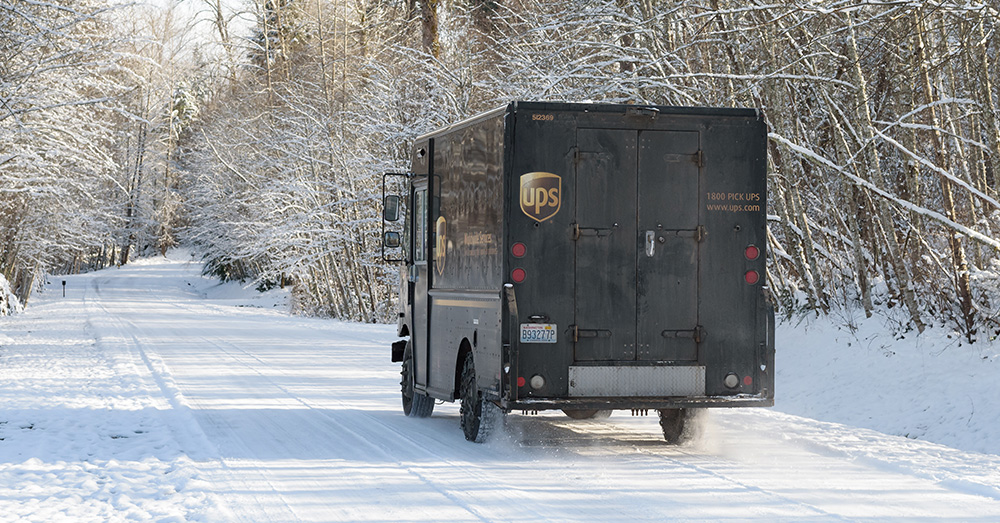 UPS delivery truck drives down a snow-covered road lined with trees.