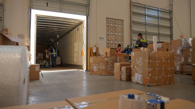 An image showing a warehouse docking bay. Goods are transferred from the back of a truck into a warehouse.
