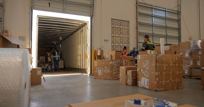 An image showing a warehouse docking bay. Goods are transferred from the back of a truck into a warehouse.