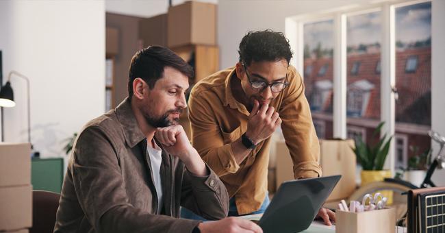 Two men look thoughtfully at a laptop screen in an office room. There are many boxes in the background.