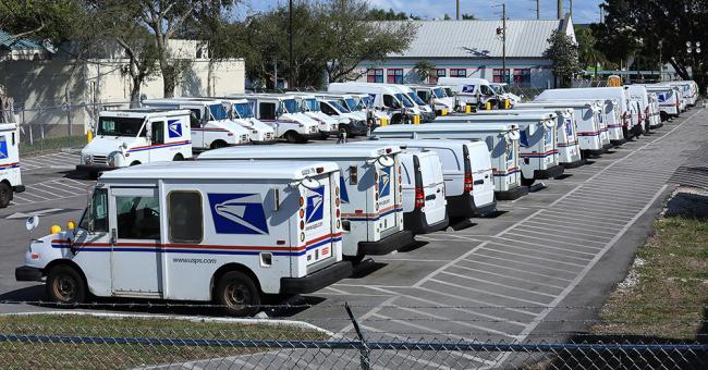 Rows of USPS delivery vehicles, including mail trucks and cargo vans, are parked in a large postal facility lot, lined up in marked spaces and ready for daily mail and package delivery.