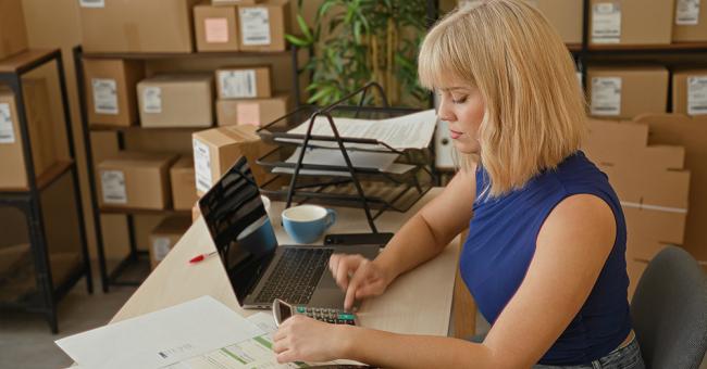 Woman using a calculator beside a laptop and paperwork, with shipping boxes stacked on shelves behind her.